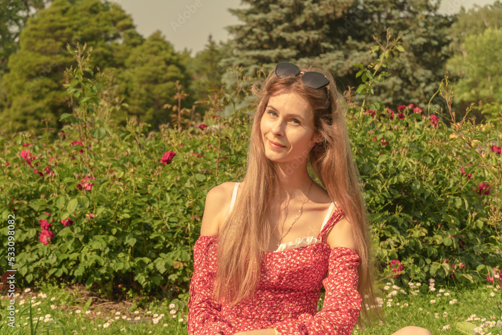 A young beautiful girl in a light red dress in a blooming summer park. Photo of attractive young charming happy woman smile good mood enjoy weekend outdoors outside park street A young beautiful girl in a light red dress in a blooming summer park. Photo of attractive young charming happy woman smile good mood enjoy weekend outdoors outside park street