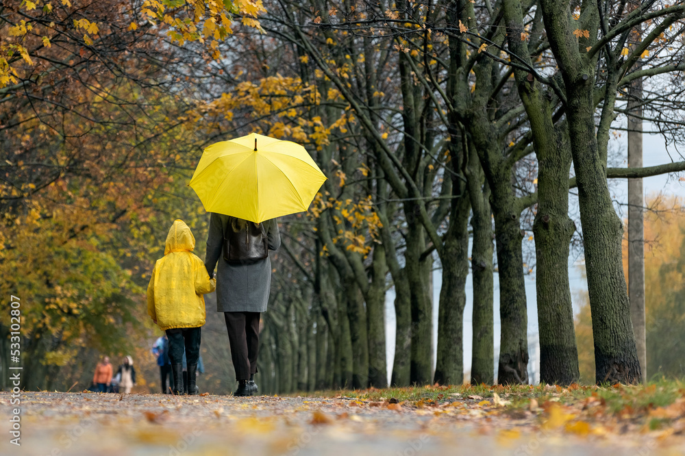 Mom and child in raincoat are walking under yellow umbrella in autumn park. Back view