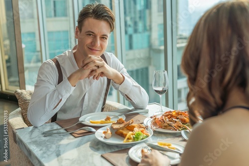 Photo of a young handsome caucasian white male guy having a good time with his asian girlfriend eating lunch in a rooftop restaurant and lounge with nice view of the city skyline