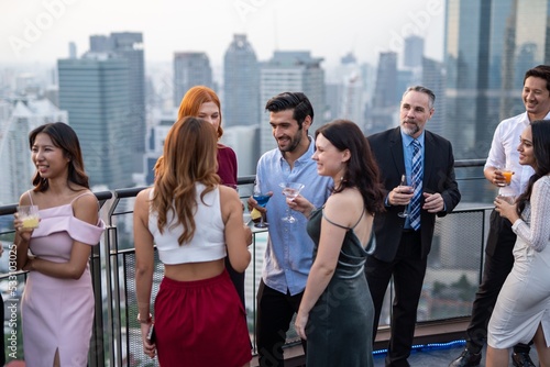 Photo of a group of businessmen and businesswomen mingle with each other drinking wine and champagne on a rooftop bar celebrating the successful of company latest deal and hit target goal of the year