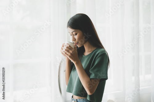 Young asian woman standing beside window and holding mug in bedroom at home, She drinking milk after wake up in the morning