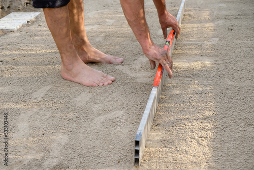 A barefoot man levels the sandy platform with an aluminum bar in his yard.