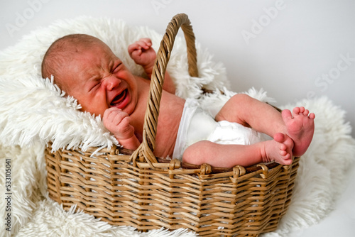 Upset new born baby in wicker basket with white fluffy plaid lying and crying.