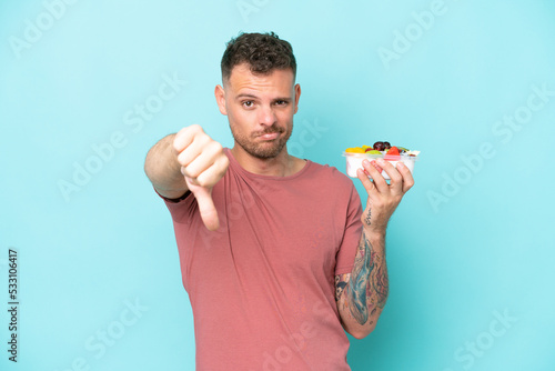 Young caucasian man holding a bowl of fruit isolated on blue background showing thumb down with negative expression