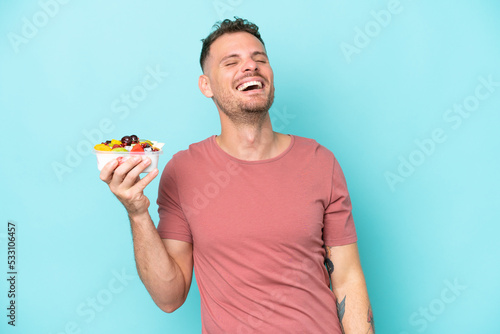 Young caucasian man holding a bowl of fruit isolated on blue background laughing