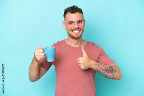 Young caucasian man holding cup of coffee isolated on blue background with thumbs up because something good has happened