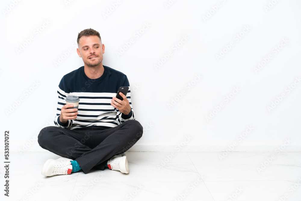 Young man sitting on the floor isolated on white background holding coffee to take away and a mobile while thinking something