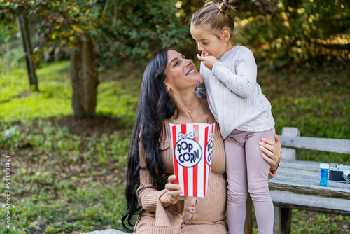 A pregnant woman and her daughter are sitting on a bench in an autumn park and eating popcorn, having fun together.