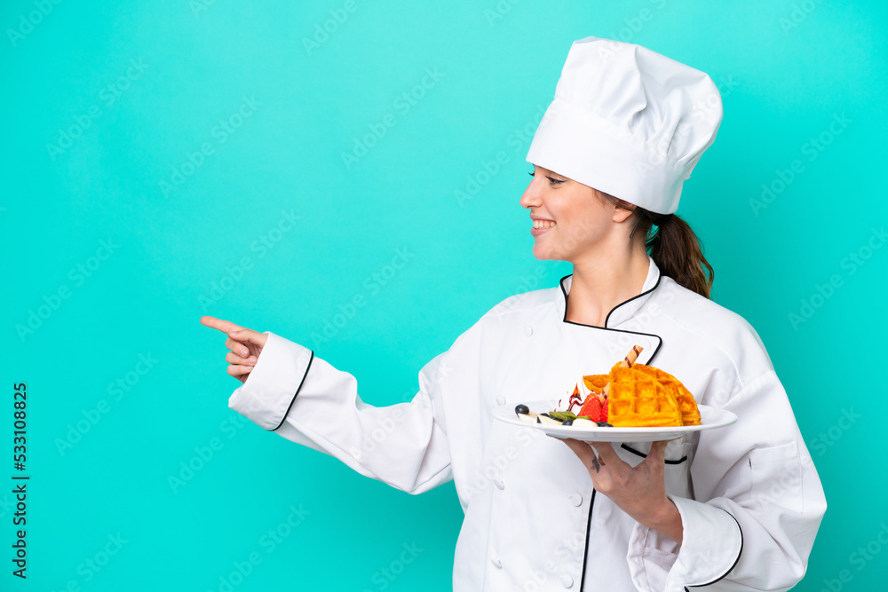 Young caucasian chef woman holding waffles isolated on blue background pointing to the side to present a product Young caucasian chef woman holding waffles isolated on blue background pointing to the side to present a product