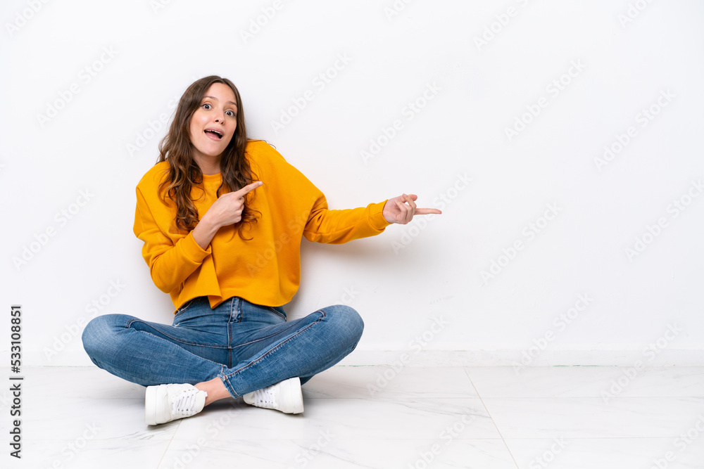 Young caucasian woman sitting on the floor isolated on white wall surprised and pointing side Young caucasian woman sitting on the floor isolated on white wall surprised and pointing side