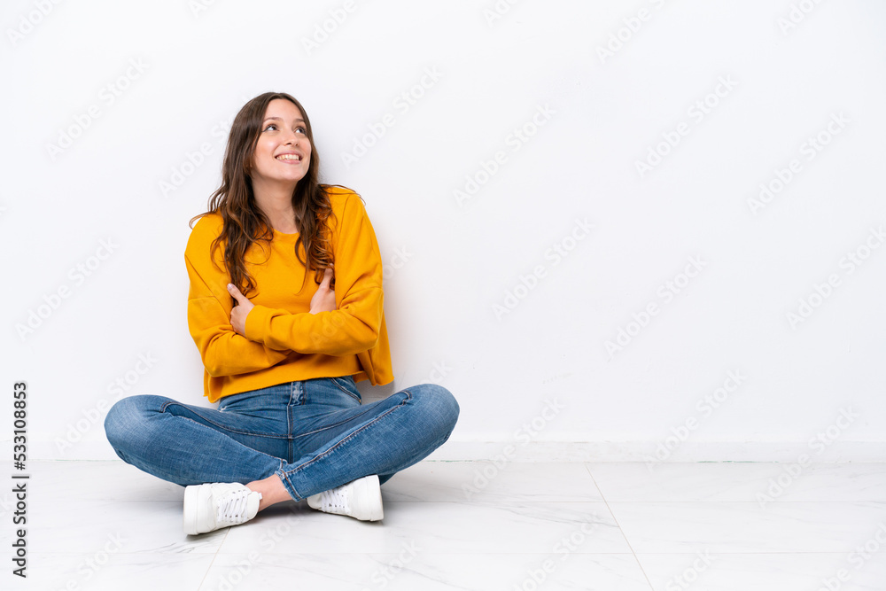 Young caucasian woman sitting on the floor isolated on white wall looking up while smiling Young caucasian woman sitting on the floor isolated on white wall looking up while smiling