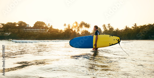 Teen boy with blue and yellow surfboard entering the waves for surfing with sunset rays on palm trees grove. Happy childhood and active sporty people vacation time concept. photo