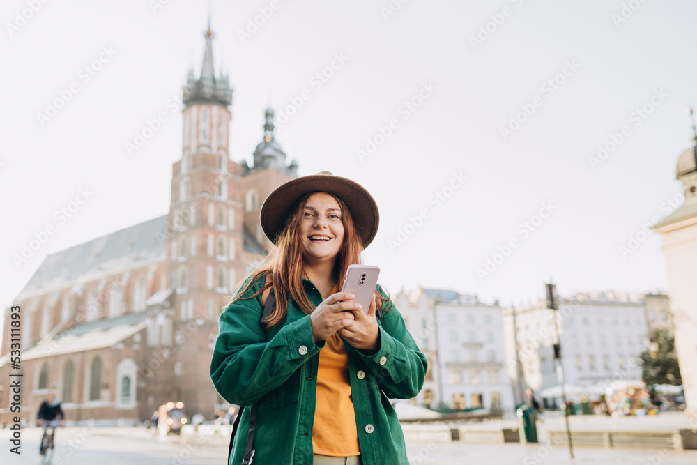 Beautiful stylish woman in hat walking on Market Square in Krakow on autumn day and holding mobile phone. Phone Communication. Urban lifestyle concept. Check social networks, send sms.