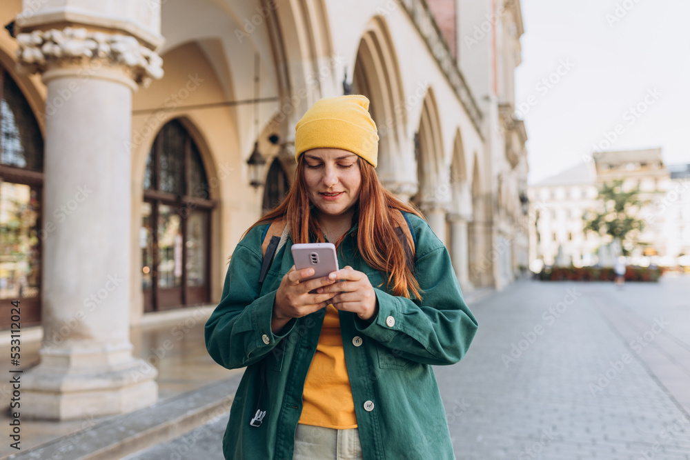 Beautiful stylish woman in hat walking on Market Square in Krakow on autumn day and holding mobile phone. Phone Communication. Urban lifestyle concept. Check social networks, send sms. Beautiful stylish woman in hat walking on Market Square in Krakow on autumn day and holding mobile phone. Phone Communication. Urban lifestyle concept. Check social networks, send sms.
