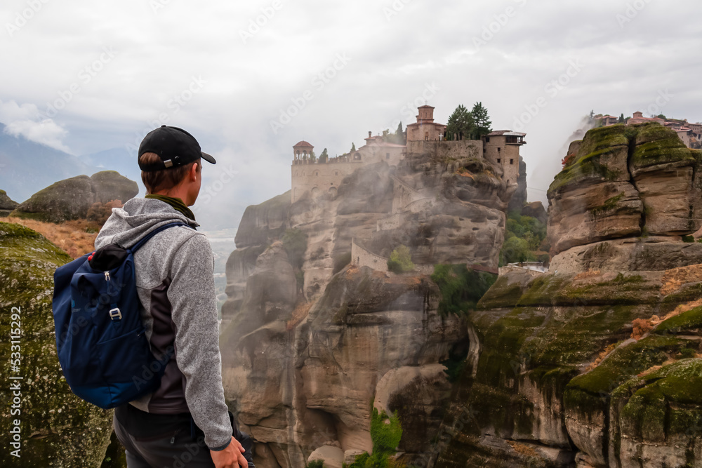 Man with scenic view of Holy Monastery of Great Meteoron appearing from fog, Kalambaka, Meteora, Thessaly, Greece, Europe. Dramatic landscape, moody vibes. Landmark build on unique rock formations Man with scenic view of Holy Monastery of Great Meteoron appearing from fog, Kalambaka, Meteora, Thessaly, Greece, Europe. Dramatic landscape, moody vibes. Landmark build on unique rock formations