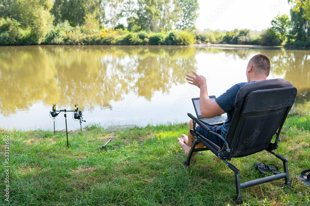 A man sits in a chair near the autumn lake and works on a laptop freelance camping. Robot out of the office, phoned with colleagues. A man sits in a chair near the autumn lake and works on a laptop freelance camping. Robot out of the office, phoned with colleagues.