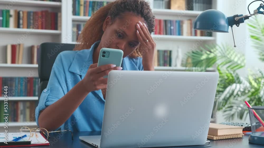 Young excited ethnic African American woman reads news on phone and rubs forehead in frustration with hand when learns about tightening of magration punishment sits in office with laptop on table Young excited ethnic African American woman reads news on phone and rubs forehead in frustration with hand when learns about tightening of magration punishment sits in office with laptop on table
