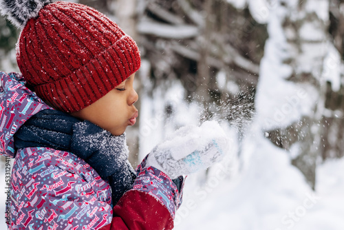 Happy little African-American girl in a red hat and overalls blowing snow off the hand.Winter, Christmas and Happy New Year concept.