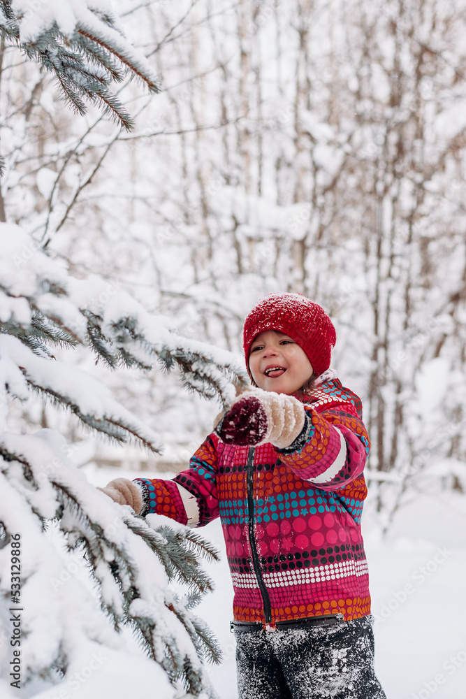 Happy little girl in a red hat and jumpsuit walks in the winter forest.Beautiful trees are covered with white snow.Winter fun,active lifestyle concept.