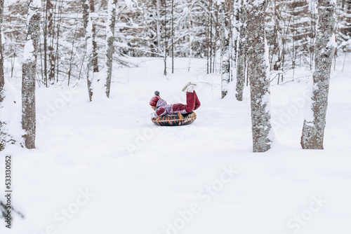 Happy little African-American girl in a red hat and jumpsuit rides on tubing in the winter park.Beautiful trees are covered with white snow.Winter fun concept.Selective focus,copy space.