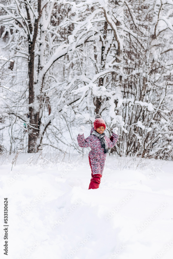 Happy little African-American girl in a red hat and jumpsuit walks in the winter forest.Beautiful trees are covered with white snow.Winter fun,active lifestyle concept.Selective focus,copy space.