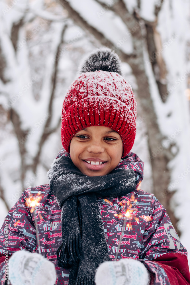 Happy little African-American girl in a red hat and jumpsuit holds a burning sparklers in her hands in a winter forest.Winter fun,merry Christmas and Happy New Year concept. Happy little African-American girl in a red hat and jumpsuit holds a burning sparklers in her hands in a winter forest.Winter fun,merry Christmas and Happy New Year concept.