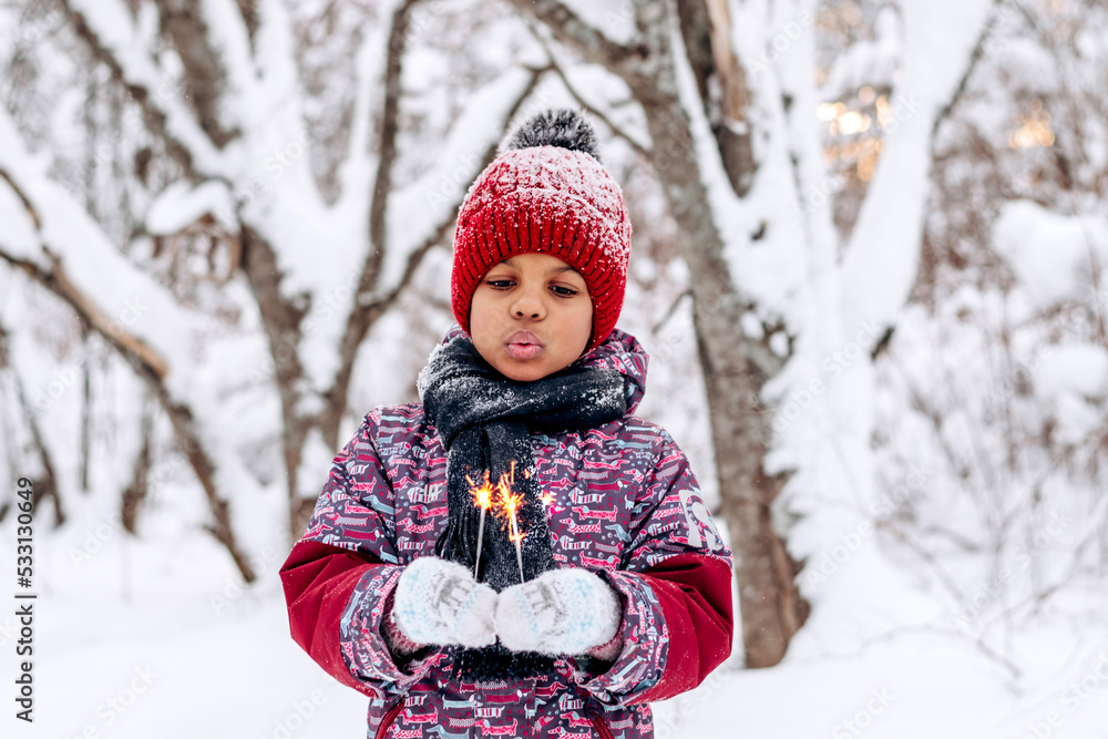 Little sad African-American girl in a red hat and jumpsuit holds a burning sparklers in her hands in a winter forest.Winter fun,merry Christmas and Happy New Year concept.