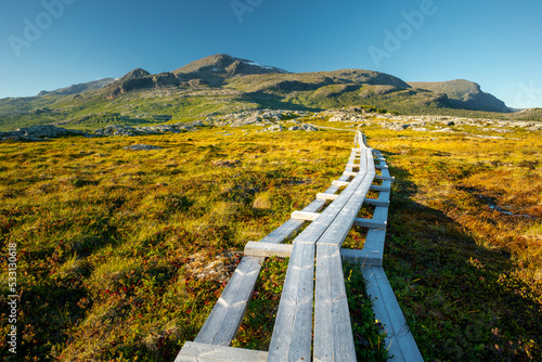 Wooden walk leading across marshlands to Nieras massive in the Stora Sjofallet National Park, Sweden. Golden hour in Swedish Arctic on a beautiful sunny day of summer. Hiking in Lapland.