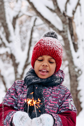 Happy little African-American girl in a red hat and jumpsuit holds a burning sparklers in her hands in a winter forest.Winter fun,merry Christmas and Happy New Year concept.