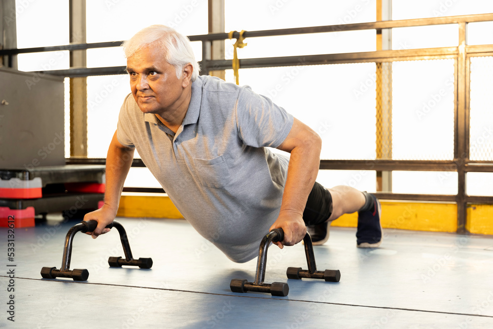 Obraz premium Active old man in sportswear doing push ups with bars at gym 