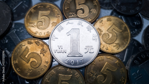 One Chinese yuan resting on several five jiao coins is shot close-up. Many coins in denominations of one Chinese yuan in the background. One yuan coin minted in 2017