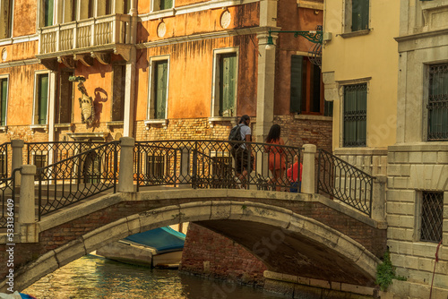 Tourists on the bridge over the canal street in Venice, old walls of houses with windows on the Venetian street in the morning sunlight, stone bridge over the canal of the city of Venice © Александр Бочкала