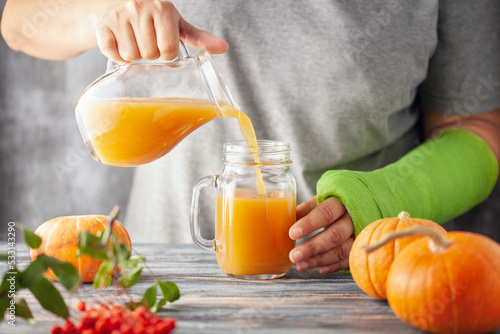 Woman with green plaster pours pumpkin juice into a glass. Girl has a broken hand wearing