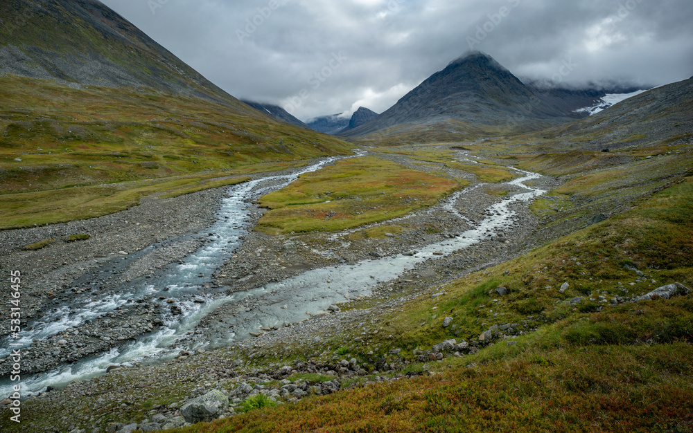 Fototapeta premium Wild river flowing through harsh, remote arctic valley on a cloudy, rainy day of summer. Basstavagge pass in Sarek National Park, Lapland, Sweden. Hiking in remote arctic wilderness of Laponia.