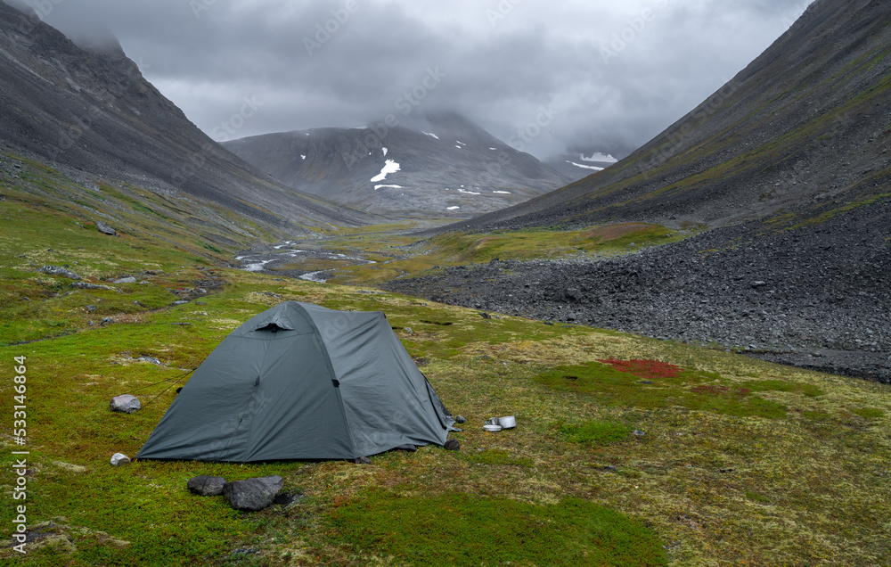 Green outdoor tent pitched in harsh, remote arctic valley on a cloudy, rainy day of summer. Basstavagge pass in Sarek National Park, Lapland, Sweden. Hiking in remote wilderness of Laponia.