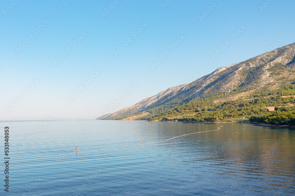 Two people are swimming in the sea on a stand up paddle against the backdrop of mountains.