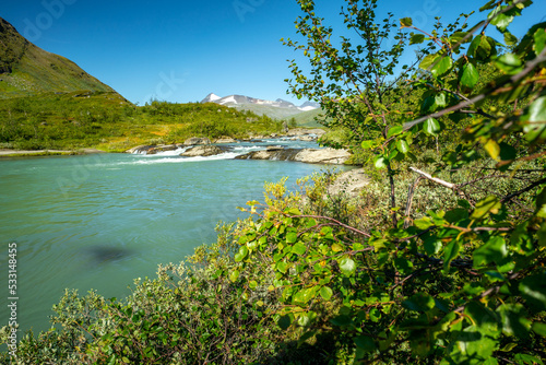 Wild glacial river flows through remote, green Arctic landscape on a sunny day of summer. Njoatsosjahka river and Ryggasberget mountain on the horizon in Sarek National Park, Sweden.