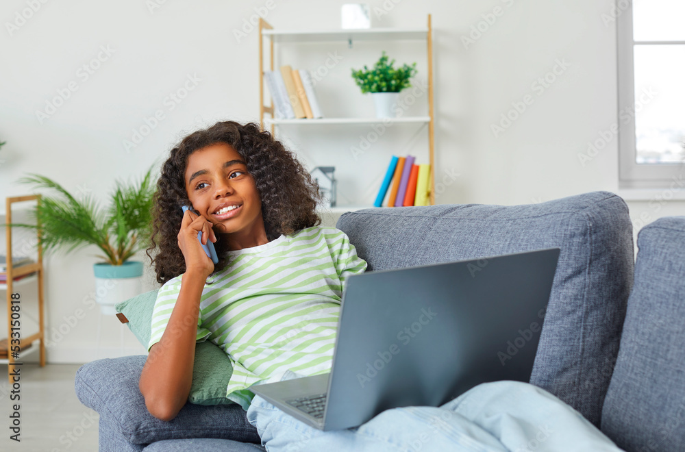 Fototapeta premium Dark-skinned teenage girl is having fun talking on phone and using laptop at same time. African American girl uses modern wireless technology while sitting on sofa at home in living room.
