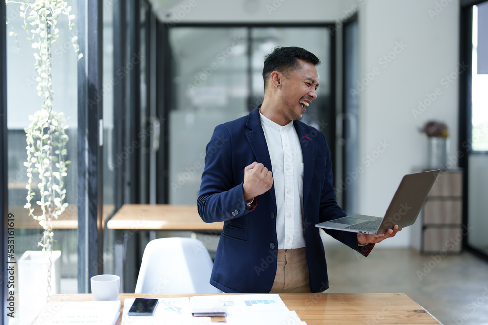 Portrait of an Asian male business owner standing with a computer showing happiness after a successful investment Portrait of an Asian male business owner standing with a computer showing happiness after a successful investment