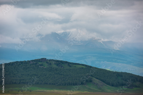 Dramatic landscape with green forest hill with view to sunlit high snowy mountain range in rainy low clouds. Atmospheric scenery with coniferous forest and large snow mountains at changeable weather.