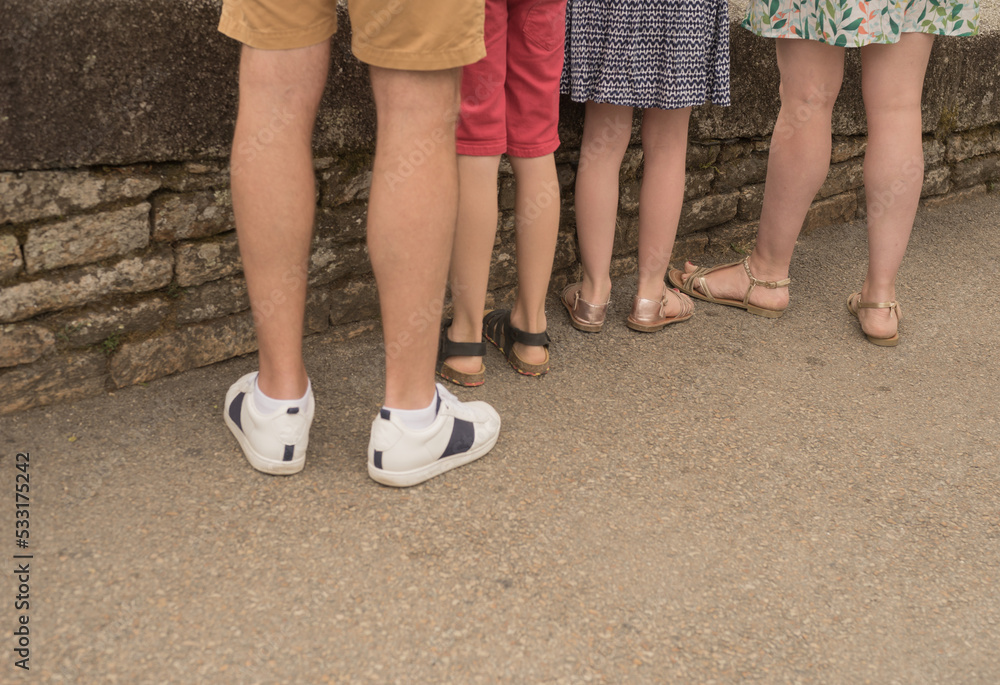 Naked tourist legs with shoes and sandals in front of a wall on asphalt in summer rear view