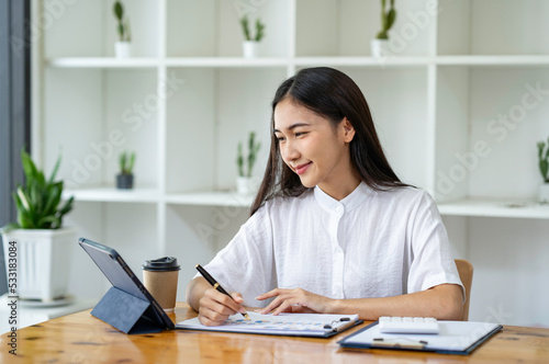 An Asian businesswoman wipes a to-do list on a tablet and responds to customer chats at a desk in a modern office.