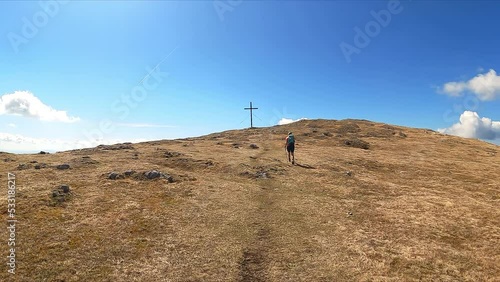 Rear view of active woman on a hiking trail arriving to the summit cross of mountain peak Foelzkogel in Hochschwab Alps, Upper Styria, Austria, Europe. Path on barren grass terrain with clear blue sky photo