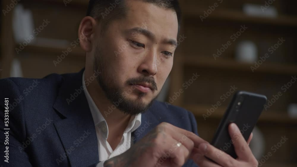Close up of a tattooed Asian businessman browsing on smart phone thinking negative on opportunity Close up of a tattooed Asian businessman browsing on smart phone thinking negative on opportunity