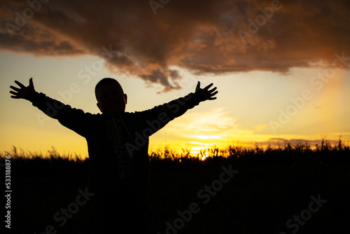 silhouette of a boy with outstretched arms in a field of clover. In the background is a sunset with beautiful colors in the sky