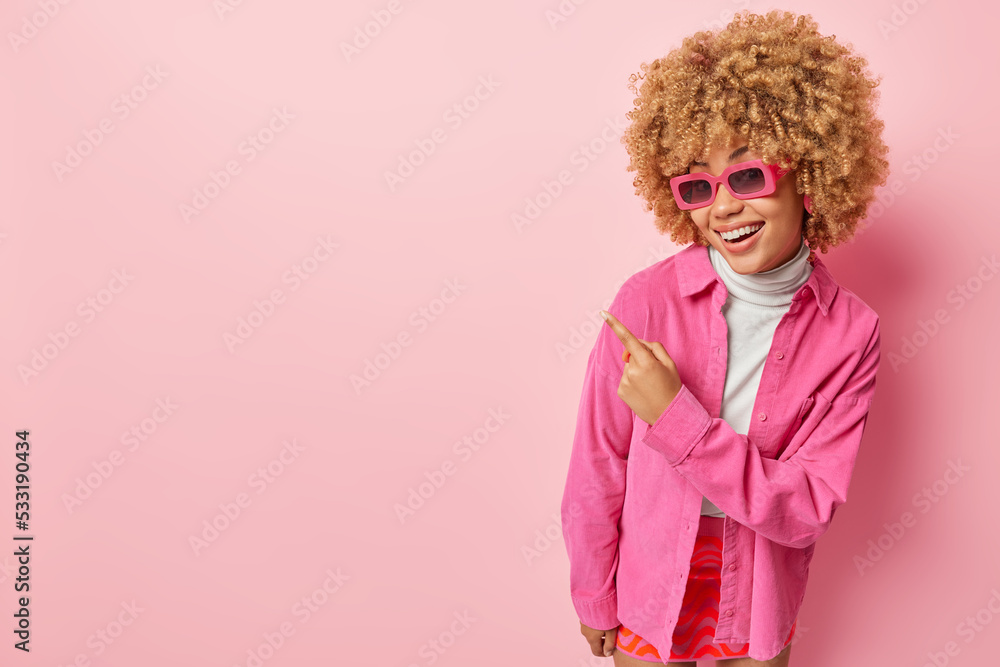 Pretty curly haired woman smiles gladfully wears sunglasses and shirt points index finger on blank space advertises product isolated over pink background. Click on this link and follow this direction