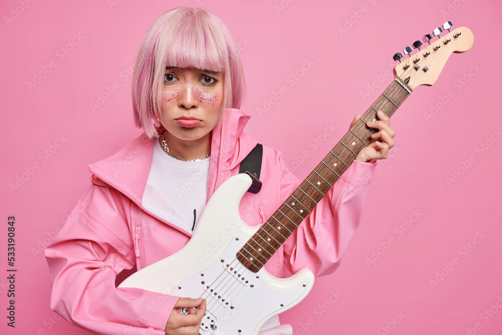 Fototapeta premium Dissatisfied Asian woman with pink dyed hair plays electic acoustic guitar learns playing musical instrument dressed in jacket frowns face has bad mood isolated over rosy background. Upset musician