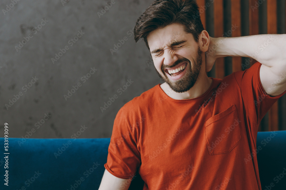 Young unhappy man wears red t-shirt feel bad suffer from pain spasm neck shoulder sit on blue sofa couch stay at home flat spend free spare time in living room indoors grey wall People lounge concept