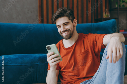 Young man wears red t-shirt hold in hand use mobile cell phone sit on blue sofa couch stay at home hotel flat rest relax spend free spare time in living room indoors grey wall. People lounge concept.