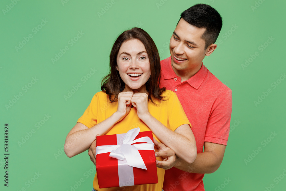 Young surprised happy couple two friends family man woman wear basic t-shirts together hold give red present box with gift ribbon bow isolated on pastel plain light green background studio portrait. Young surprised happy couple two friends family man woman wear basic t-shirts together hold give red present box with gift ribbon bow isolated on pastel plain light green background studio portrait.